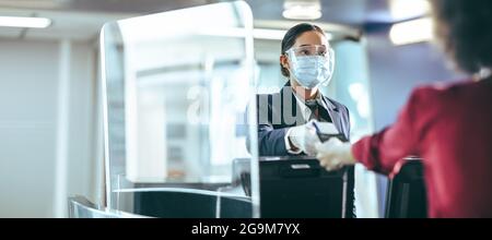 L'assistente di volo indossa una maschera protettiva e una visiera protettiva che assiste il passeggero all'aeroporto. Check-in al banco del dipendente che dà la carta d'imbarco al travel Foto Stock