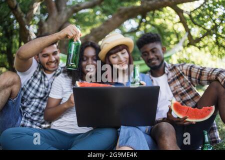 Gruppo di quattro persone multietniche che utilizzano un computer portatile wireless per le videochiamate all'aperto. Felici gli amici che si godono il tempo di picnic con tecnologia moderna, cibo e bevande. Foto Stock