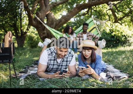 L'uomo indiano e la donna caucasica riposano su morbida coperta all'aperto con smartphone moderni in mani. Coppia afro-americana seduta in amaca e bevendo birra dietro. Quattro amici diversi al picnic. Foto Stock