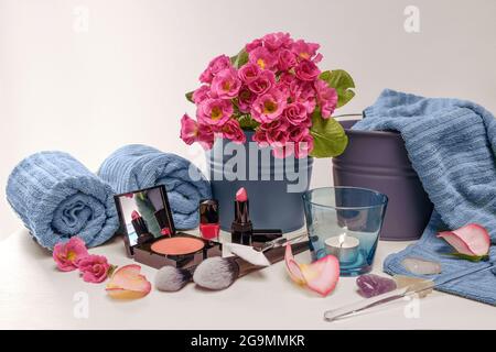 Make-up e utensili di bellezza come polvere per il viso, rossetto e smalto per unghie con spazzole cosmetiche, asciugamani blu, candela e fiori rosa in vaso, sfondo chiaro Foto Stock