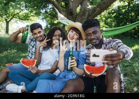 Gruppo allegro di quattro amici diversi seduti nel giardino verde con cocomero e birra in mani e prendendo selfie su smartphone moderno, concetto di persone, tecnologia e tempo libero. Foto Stock