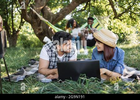 Giovane ragazzo indiano e donna caucasica sdraiati su una morbida plaid in giardino e utilizzando un computer portatile wireless. Coppia afroamericana che si rilassa in amaca con birra in mano. Quattro amici felici al picnic. Foto Stock