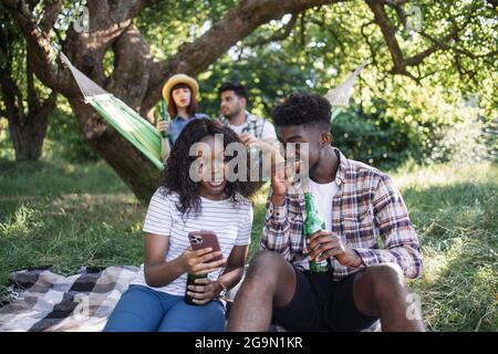 Coppia afroamericana che usa uno smartphone moderno mentre si siede insieme sulla plaid nel giardino estivo. Amici multirazziali che chiacchierano e bevono birra in amaca sullo sfondo. Foto Stock