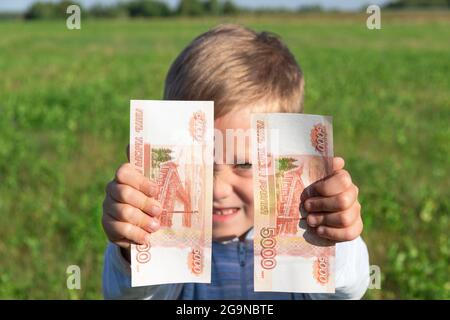 Un bambino soddisfatto, un ragazzo preschooler, tiene i rubli di carta nelle sue mani all'aperto sullo sfondo di un campo con erba verde in un'estate luminosa e soleggiata Foto Stock