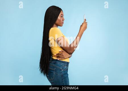 Vista laterale di Serious Black Female con smartphone su sfondo blu Foto Stock