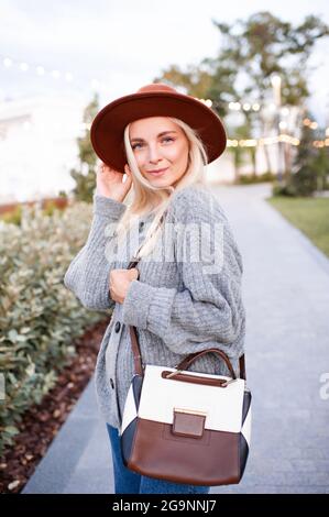 Felice elegante ragazza bionda 19-20 anni indossare cappello, maglia maglia maglia in lana jumper e tenere borsa in pelle camminare nel parco all'aperto. Guardando la fotocamera. Felicità. AUT Foto Stock