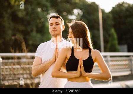 Giovane coppia uomo e donna meditare in piedi, yoga sul prato della città, estate sera, stile di vita Foto Stock