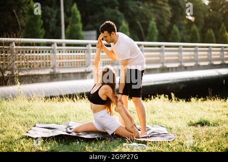 Personal trainer uomo istruire giovane donna, yoga sul prato della città, estate sera, in forma Foto Stock