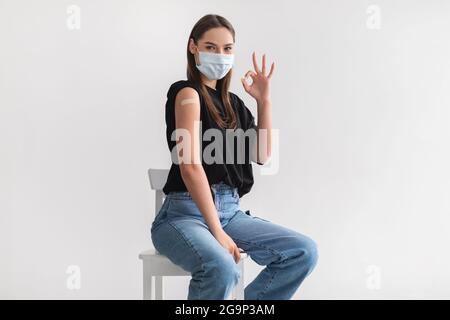 Giovane donna caucasica in maschera facciale dopo immunizzazione covid-19, mostrando spalla con aiuto di banda, gesturing OK Foto Stock