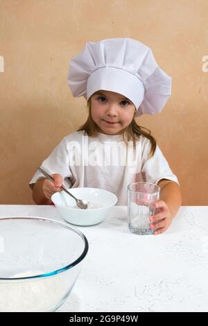 Una bambina vestita come cuoco impasta l'impasto. Cucina bambino stile di vita concetto. Il capretto ama, ha divertimento, studia e gioca nella cucina Foto Stock