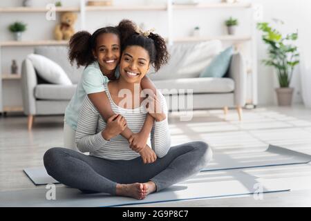 Madre e figlia nere sportive sedute sul tappetino fitness, abbraccianti Foto Stock