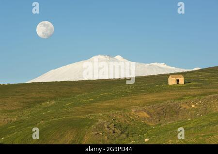 Pendio verde con un baracchio e l'Etna coperto di neve sotto la luna piena nel cielo blu della sera Foto Stock