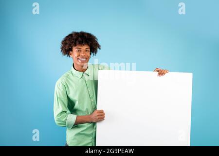 Offerta speciale. Felice ragazzo nero teen che tiene vuoto bordo vuoto, mostrandolo a macchina fotografica sopra sfondo blu studio Foto Stock