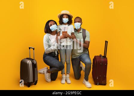 Viaggi sicuri. Famiglia afroamericana che indossa maschere mediche, in posa con valigie e passaporti su sfondo giallo Foto Stock