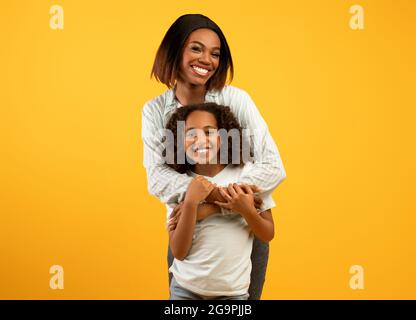 Amore della madre. Felice amorevole mamma afroamericana coccolando la figlia dalla parte posteriore, in piedi su sfondo giallo Foto Stock