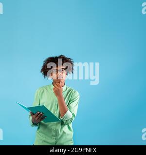 Premuroso ragazzo teen afro-americano in occhiali che tengono il notebook e brocciare il mento, guardando in su allo spazio libero, raccolto Foto Stock