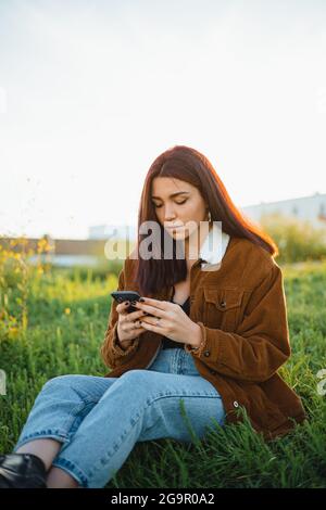 Un adolescente che invia un messaggio di testo ai suoi amici sul suo telefono mentre si siede su un prato verde durante il tramonto Foto Stock