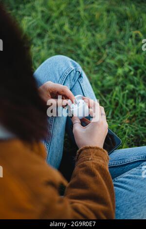 Una donna che apre la custodia di ricarica degli auricolari wireless bianchi mentre si siede sull'erba (verticale) Foto Stock