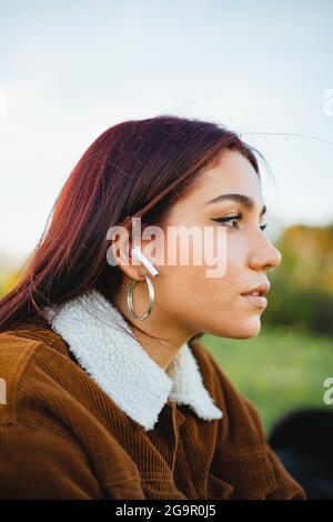 Una ragazza adolescente che ascolta la musica attraverso auricolari wireless bianchi. È seduta sull'erba durante il tramonto. Foto Stock