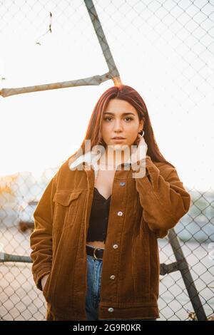 Ritratto di bella ragazza teenager in piedi contro recinzione catena-link guardando la macchina fotografica e toccando il collo durante il tramonto. Indossa un j marrone Foto Stock