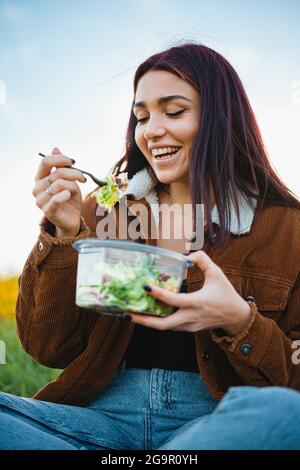 Ridendo ragazza adolescente godendo di mangiare un'insalata. È sul campo durante il tramonto. Foto Stock