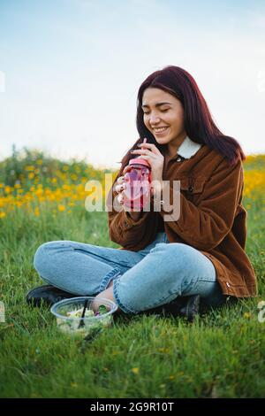 Ridendo la ragazza adolescente che beve acqua in un vaso rosso seduto sull'erba di una collina. Ha un'insalata accanto a lei. Foto Stock