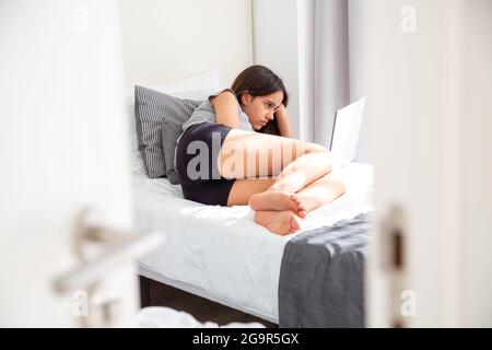 Concentrato ragazza teen usando il suo laptop sdraiato a letto. Vista dalla porta aperta a metà Foto Stock