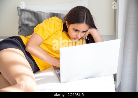 Teen ragazza concentrata con una camicia gialla utilizzando il suo computer portatile sdraiato a letto Foto Stock
