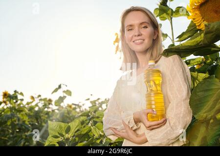 Giovane donna bionda sorridente in abito bianco che tiene una bottiglia di olio di girasole in piedi contro grandi fiori di fronte telecamera sul campo Foto Stock
