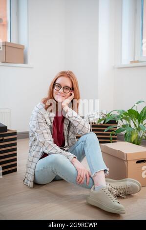 In un nuovo appartamento a luce bianca, una ragazza si siede sul pavimento e si pone sullo sfondo di scatole di cartone e un fiore in vaso. Foto Stock