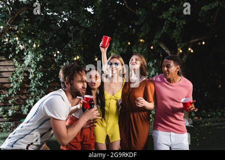 Uomo che canta karaoke vicino amici interracial con tazze di plastica all'aperto Foto Stock