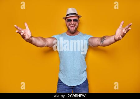 Avvicinati, mi abbraccio. Studio shot di positivo amichevole bearded uomo in abito trendy e cappello che tira le mani verso la macchina fotografica, invitando i compagni a. Foto Stock