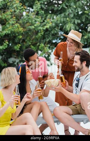 uomo sorridente che tiene ciotola di popcorn vicino allegri amici interrazziali che bevono birra Foto Stock