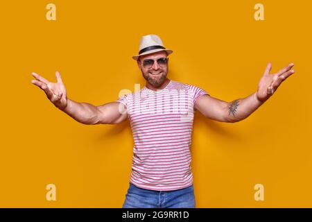 Avvicinati, mi abbraccio. Studio shot di positivo amichevole bearded uomo in trendy spogliato vestito e cappello che tira le mani verso la macchina fotografica, invitante Foto Stock