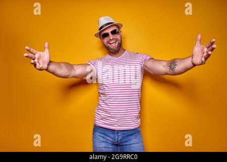 Avvicinati, mi abbraccio. Studio shot di positivo amichevole bearded uomo in trendy spogliato vestito e cappello che tira le mani verso la macchina fotografica, invitante Foto Stock