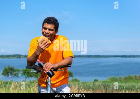 Uomo affamato che mangia un hamburger e beve la cola da una bottiglia in bicicletta Foto Stock