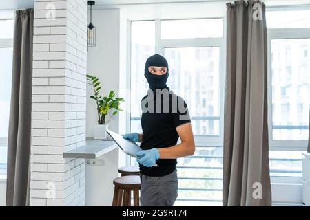 Ladro con balaclava nera rubare laptop. Il burglar commette un crimine in appartamento di lusso Foto Stock