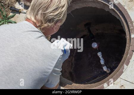 L'uomo si è piegato sopra il pozzo dell'acqua e registra le letture del contatore dell'acqua. Controllo e fissaggio del contatore. Foto Stock