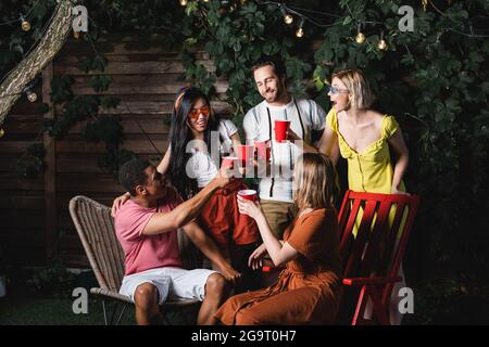 Felici amici multiculturali che tostano con tazze di plastica sotto la terraferma all'aperto Foto Stock