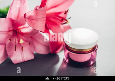 Vasetto con crema idratante circondato da fiori di giglio rosa su sfondo grigio. Cosmetici biologici. Cura della pelle Foto Stock