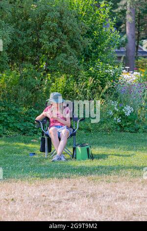 donna anziana con un cappello seduto su una sedia pieghevole nel parco che tiene uno smartphone e ascolta musica. White Rock, British Columbia, Canada, luglio 2,2021. Via vie Foto Stock