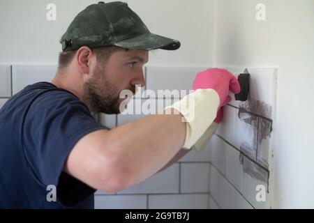 Il lavoratore applica la grota grigia a piastrelle bianche con cazzuola di gomma. Foto Stock