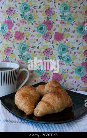 Tre croissant al burro su un piatto nero e una grande tazza di caffè nero, piccola colazione in stile vecchio stile con sfondo floreale Foto Stock