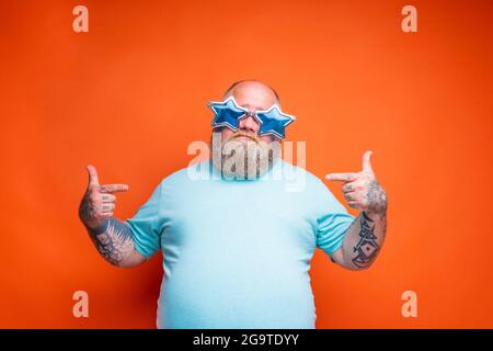 L'uomo del dubbio grasso con la barba, i tatuaggi e gli occhiali da sole è incerto per qualcosa Foto Stock