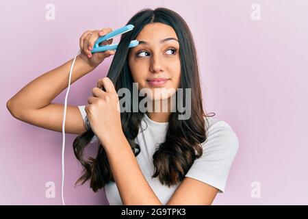 Giovane donna ispanica che tiene la piastra per capelli sorridente guardando al lato e fissando via pensare. Foto Stock