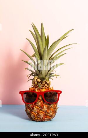 Concetto estivo di ananas divertente con occhiali da sole su assi di legno blu con sfondo rosa. Foto Stock