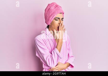 Giovane uomo che indossa donna si compongono portando asciugamano da doccia sulla testa e accappatoio annoiato yawning stanco coprire la bocca con la mano. Riposante e sonnolenza. Foto Stock