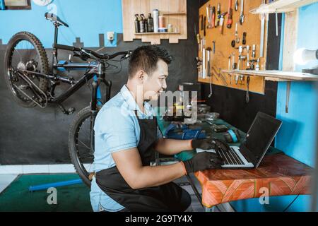 un meccanico di bicicletta in un grembiule che indossa guanti seduti mentre si utilizza un computer portatile per cercare i pezzi di ricambio Foto Stock