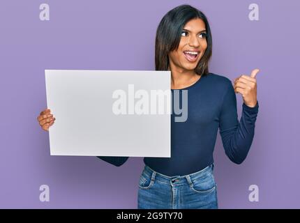 Giovane donna transessuale latina che tiene vuoto banner che punta il pollice verso il lato sorridendo felice con la bocca aperta Foto Stock