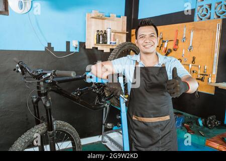 bel meccanico di bicicletta in grembiule sorridente indossando guanti e tenendo la chiave a bussola con i pollici verso l'alto Foto Stock